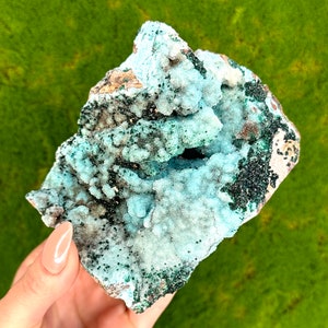 May include: A hand holds a piece of raw mineral with a light blue, crystalline structure. The mineral has areas of dark green and brown, with a rough, uneven surface. The background is a solid, bright green.