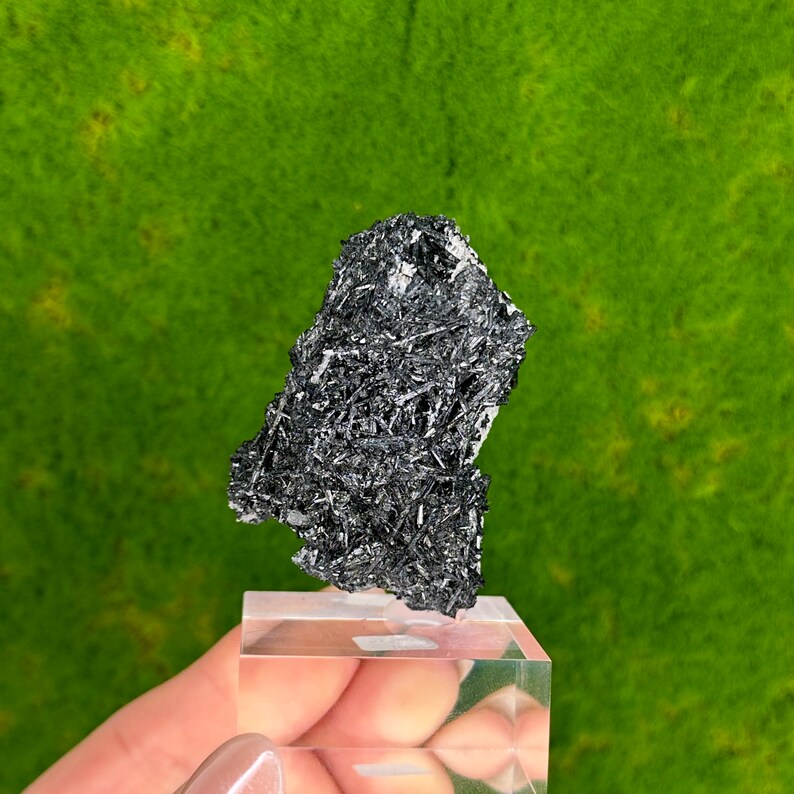 May include: A black, naturally formed, crystal cluster with a rough, textured surface. The crystal is displayed on a clear acrylic stand.