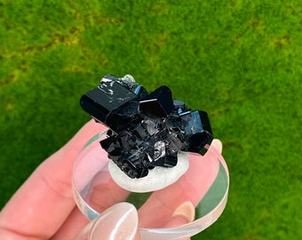 May include: A cluster of black crystals on a clear display stand. The crystals have a sharp, geometric shape and a glossy finish.