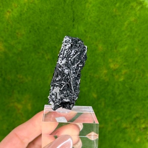 May include: A black and white crystal with a fibrous texture. The crystal is displayed on a clear acrylic stand.
