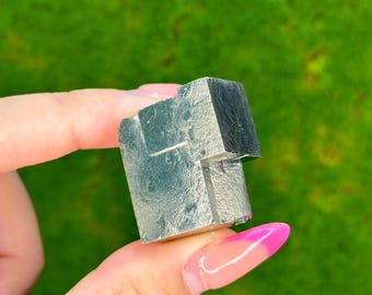 Può includere: Un cristallo di pirite grigio metallico con forma cubica. Il cristallo ha una superficie ruvida e testurizzata ed è un minerale naturale.