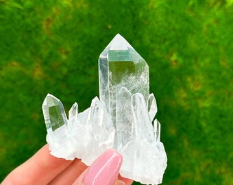 May include: A cluster of clear quartz crystals with a large, pointed crystal in the center. The crystals are arranged in a cluster, with some of the crystals pointing upwards and others pointing downwards.