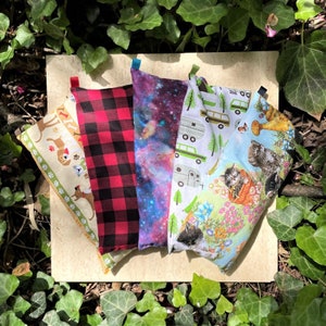 May include: Five fabric eye pillows, each with a unique design. The pillows are arranged in a row on a light brown surface. The designs include a plaid pattern, a galaxy print, a camper van print, a floral print with cats, and a dog print.