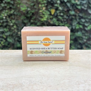 Pu&ograve; includere: Una saponetta rettangolare di burro di karit&egrave; rosa con un'etichetta gialla, arancione e bianca che recita "The Soap Opera" e "SCENTED SHEA BUTTER SOAP Coconut" con un numero di telefono e un indirizzo di sito web.
