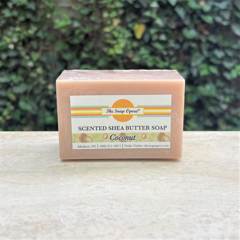 Pu&ograve; includere: Una saponetta rettangolare di burro di karit&egrave; rosa con un'etichetta gialla, arancione e bianca che recita "The Soap Opera" e "SCENTED SHEA BUTTER SOAP Coconut" con un numero di telefono e un indirizzo di sito web.