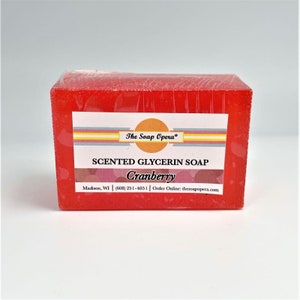 Pu&ograve; includere: Una saponetta rossa di glicerina profumata al mirtillo rosso con un'etichetta bianca che recita "The Soap Opera" e "SCENTED GLYCERIN SOAP Cranberry Madison, WI | (608) 251-4051 | Order Online: thesoapopera.com"