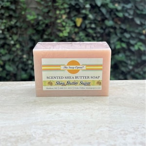 Pu&ograve; includere: Una saponetta rettangolare di burro di karit&egrave; rosa con il testo "The Soap Opera" e "SCENTED SHEA BUTTER SOAP Shea Butter Sugar" stampato sull'etichetta. L'etichetta include anche le informazioni di contatto dell'azienda: "Madison, WI (606) 251-4051 | Order Online: thesoapopera.com".