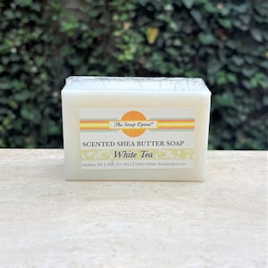 Pu&ograve; includere: Una saponetta bianca di burro di karit&egrave; profumato con il testo "The Soap Opera" e "SCENTED SHEA BUTTER SOAP White Tea" sull'etichetta. L'etichetta include anche le informazioni di contatto dell'azienda.