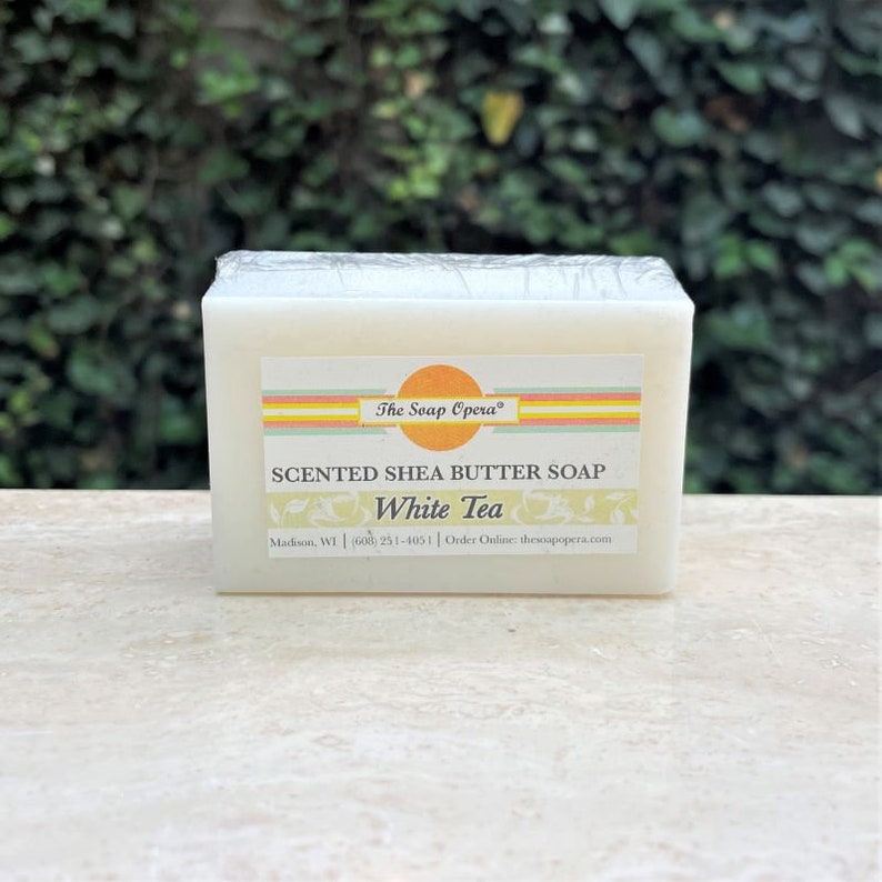 Pu&ograve; includere: Una saponetta bianca di burro di karit&egrave; profumato con il testo "The Soap Opera" e "SCENTED SHEA BUTTER SOAP White Tea" sull'etichetta. L'etichetta include anche le informazioni di contatto dell'azienda.