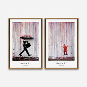 May include: Two framed prints of street art by Banksy. The first print shows a man walking in the rain with an umbrella. The second print shows a child standing in the rain with arms outstretched. Both prints have a colorful rain effect and the text "Banksy Colored Rain".