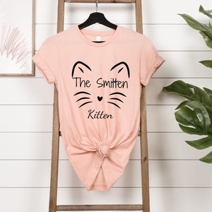 May include: A peach-colored t-shirt with a black cat face design and the words "The Smitten Kitten." The shirt is knotted at the waist and hangs on a black hanger. A wooden ladder and a framed picture are in the background.