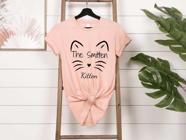 May include: A peach-colored t-shirt with a black cat face design and the words "The Smitten Kitten." The shirt is knotted at the waist and hangs on a black hanger. A wooden ladder and a framed picture are in the background.
