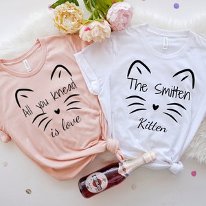 May include: Two t-shirts, one blush pink and one white, with black cat face designs. The pink shirt reads "All you knead is love," and the white shirt says "The Smitten Kitten." A bottle of sparkling wine and flowers are in the background.