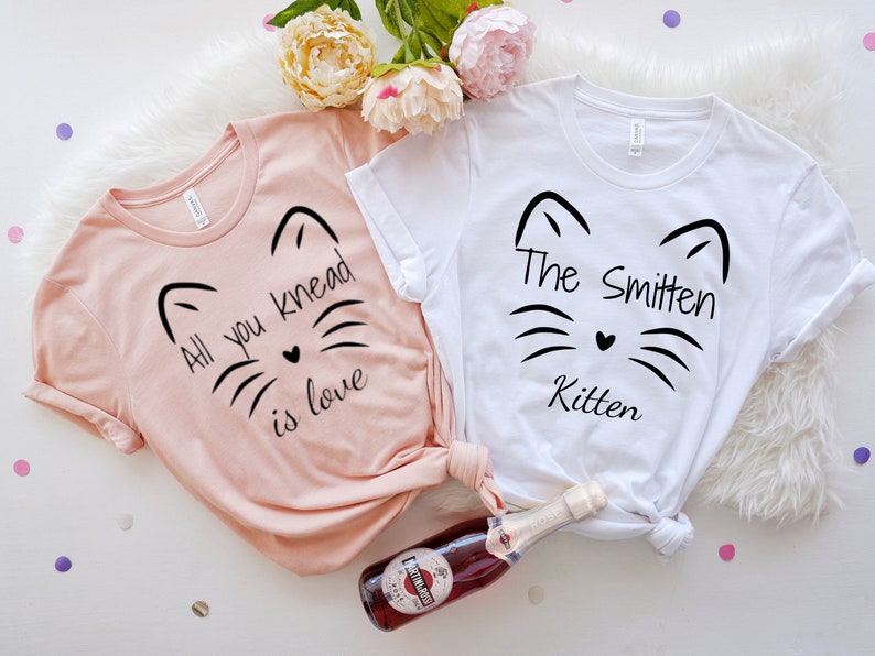 May include: Two t-shirts, one blush pink and one white, with black cat face designs. The pink shirt reads "All you knead is love," and the white shirt says "The Smitten Kitten." A bottle of sparkling wine and flowers are in the background.