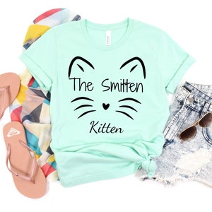 May include: A mint green t-shirt with the text "The Smitten Kitten" and a cat face design. The shirt is styled with denim shorts, sunglasses, a patterned scarf, and pink flip-flops.
