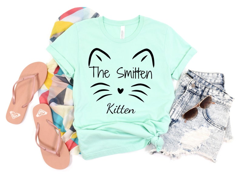 May include: A mint green t-shirt with the text "The Smitten Kitten" and a cat face design. The shirt is styled with denim shorts, sunglasses, a patterned scarf, and pink flip-flops.