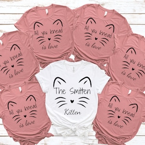 May include: A collection of pink and white t-shirts with cat face designs. The pink shirts have the text "All you knead is love." The white shirt says "The Smitten Kitten."