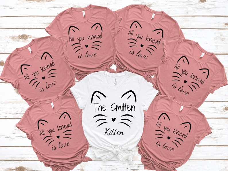 May include: A collection of pink and white t-shirts with cat face designs. The pink shirts have the text "All you knead is love." The white shirt says "The Smitten Kitten."