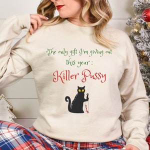 May include: Beige sweatshirt with the text "The only gift I'm giving out this year: Killer Pussy." A black cat with yellow eyes and a bloody knife is printed below the text. The sweatshirt is a festive holiday design.