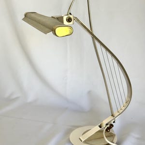 May include: A silver-toned metal desk lamp featuring a curved, harp-like frame with thin vertical wires. The lamp has a rectangular, textured shade with a yellow oval end cap and a circular base, connected by an adjustable hinge mechanism.