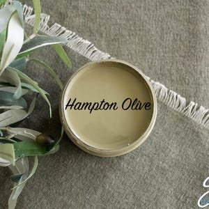 May include: A small jar of paint labeled "Hampton Olive" in a light brown color. The jar is sitting on a textured, light brown fabric surface. The text "Silk All-in-One Mineral Paint" is visible in the lower right corner of the image.