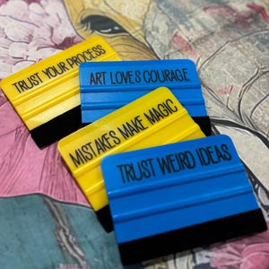 May include: Four yellow and blue squeegees with black edges, each bearing a different inspirational quote in black text. The quotes include "Trust Your Process," "Art Loves Courage," "Mistakes Make Magic," and "Trust Weird Ideas."