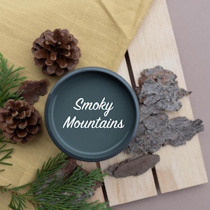 May include: A black candle jar with the text "Smoky Mountains" in white lettering. The jar is surrounded by pine cones, pine needles, and bark on a wooden surface.