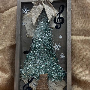 K&ouml;nnte beinhalten: Eine gerahmte Weihnachtsbaumdekoration mit einer Schleife aus Sackleinen, Notenpapier und einem Violinschl&uuml;ssel. Der Baum besteht aus gr&uuml;nen Glassplittern.
