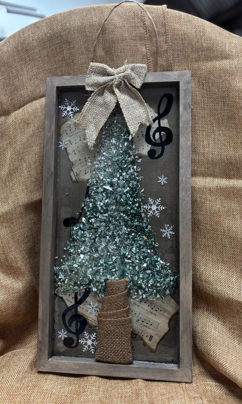 K&ouml;nnte beinhalten: Eine gerahmte Weihnachtsbaumdekoration mit einer Schleife aus Sackleinen, Notenpapier und einem Violinschl&uuml;ssel. Der Baum besteht aus gr&uuml;nen Glassplittern.