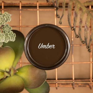 May include: A small jar of brown paint with the word "Umber" written in white on the label. The jar is sitting on a copper wire mesh surface.