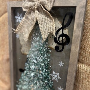 K&ouml;nnte beinhalten: Eine gerahmte Weihnachtsbaumdekoration mit einer Schleife aus Sackleinen, einem Violinschl&uuml;ssel und einem gr&uuml;n-wei&szlig;en glitzerartigen Material. Der Rahmen ist aus Holz und hat eine rustikale Optik.