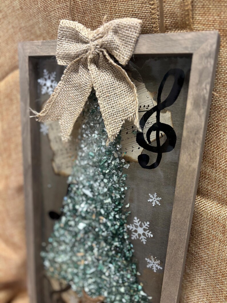 K&ouml;nnte beinhalten: Eine gerahmte Weihnachtsbaumdekoration mit einer Schleife aus Sackleinen, einem Violinschl&uuml;ssel und einem gr&uuml;n-wei&szlig;en glitzerartigen Material. Der Rahmen ist aus Holz und hat eine rustikale Optik.
