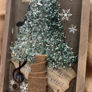 K&ouml;nnte beinhalten: Ein gerahmtes Kunstwerk mit einem Weihnachtsbaum aus gr&uuml;nen und silbernen Glassplittern. Der Baum ist von Notenbl&auml;ttern umgeben, auf denen die W&ouml;rter "Happy we're singing" und "Merry bells are" zu sehen sind. Der Baum hat einen Stamm aus Sackleinen und ist mit wei&szlig;en Schneeflocken verziert.