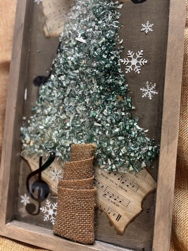 K&ouml;nnte beinhalten: Ein gerahmtes Kunstwerk mit einem Weihnachtsbaum aus gr&uuml;nen und silbernen Glassplittern. Der Baum ist von Notenbl&auml;ttern umgeben, auf denen die W&ouml;rter "Happy we're singing" und "Merry bells are" zu sehen sind. Der Baum hat einen Stamm aus Sackleinen und ist mit wei&szlig;en Schneeflocken verziert.