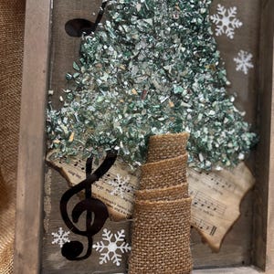 K&ouml;nnte beinhalten: Eine gerahmte Weihnachtsbaumdekoration aus gr&uuml;nen und silbernen Glassplittern. Der Baum ist mit einem Jutesockel und einem schwarzen Violinschl&uuml;ssel verziert. Der Hintergrund ist ein altes Notenblatt mit Schneeflocken.