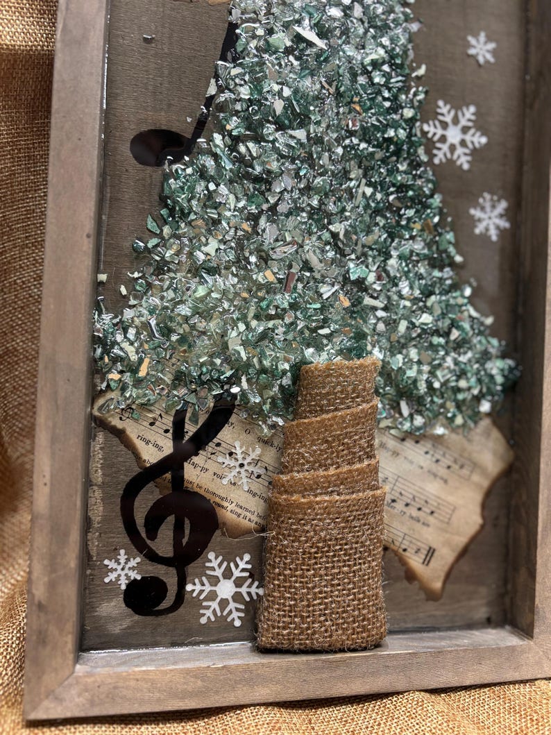 K&ouml;nnte beinhalten: Eine gerahmte Weihnachtsbaumdekoration aus gr&uuml;nen und silbernen Glassplittern. Der Baum ist mit einem Jutesockel und einem schwarzen Violinschl&uuml;ssel verziert. Der Hintergrund ist ein altes Notenblatt mit Schneeflocken.