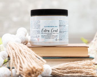 May include: A jar of Silk All-In-One Mineral Paint, labeled "Extra Coat," sits atop a stack of books. The jar has a black lid and a white label with product information. A decorative tassel and white beaded garland are in the foreground.