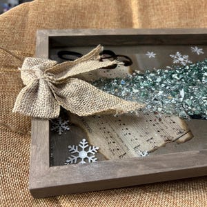 K&ouml;nnte beinhalten: Ein rustikaler Holzrahmen mit einer Schleife aus Sackleinen, einem Notenblatt und einem Weihnachtsbaum aus gr&uuml;nen Glasperlen. Der Rahmen h&auml;ngt an einem braunen Sackleinenhintergrund.