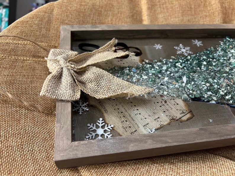 K&ouml;nnte beinhalten: Ein rustikaler Holzrahmen mit einer Schleife aus Sackleinen, einem Notenblatt und einem Weihnachtsbaum aus gr&uuml;nen Glasperlen. Der Rahmen h&auml;ngt an einem braunen Sackleinenhintergrund.