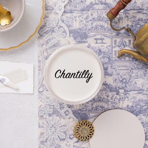 May include: An overhead shot features a white container labeled "Chantilly," alongside a gold-rimmed teacup, a small palette knife with a paint swatch, and a vintage brass teapot. The background includes a blue and white toile pattern and white lace.
