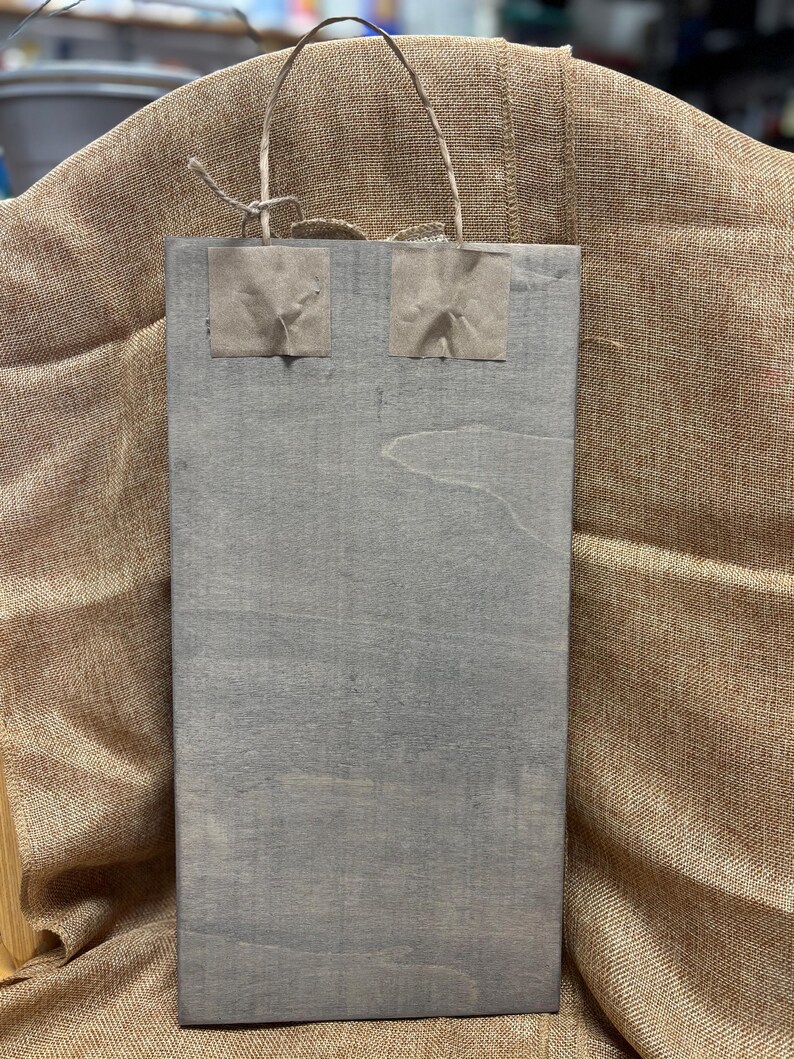 K&ouml;nnte beinhalten: Eine graue Papiert&uuml;te mit zwei braunen Papierquadraten, die auf die Vorderseite geklebt sind. Die Tasche hat einen Seilgriff.