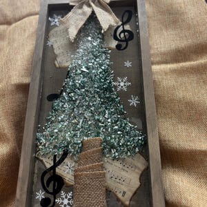 K&ouml;nnte beinhalten: Eine gerahmte Weihnachtsbaumdekoration mit einer Schleife aus Sackleinen, Noten und einem gr&uuml;nen Glitzerbaum. Der Baum besteht aus zerbrochenem Glas und ist von Notenbl&auml;ttern umgeben.