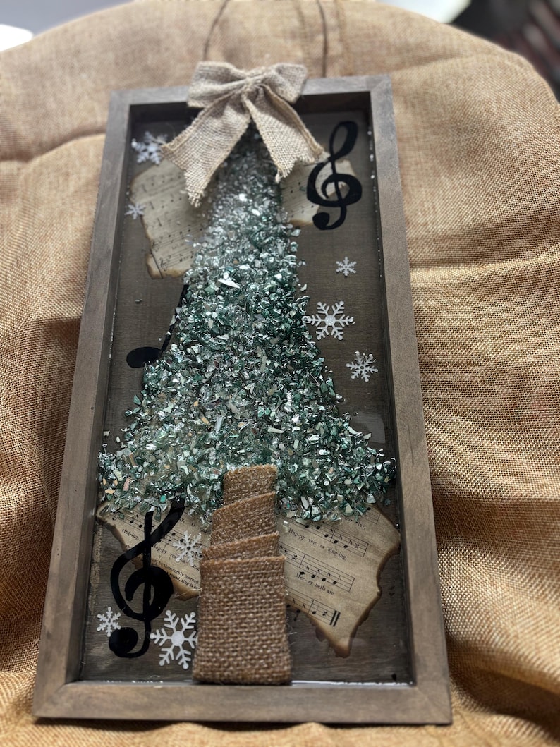 K&ouml;nnte beinhalten: Eine gerahmte Weihnachtsbaumdekoration mit einer Schleife aus Sackleinen, Noten und einem gr&uuml;nen Glitzerbaum. Der Baum besteht aus zerbrochenem Glas und ist von Notenbl&auml;ttern umgeben.