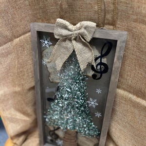 K&ouml;nnte beinhalten: Ein rustikaler Holzrahmen mit einer Schleife aus Sackleinen und einem gr&uuml;nen Glitzer-Weihnachtsbaum im Inneren. Der Rahmen hat eine Glasfront und ist mit Noten und Schneeflocken verziert.
