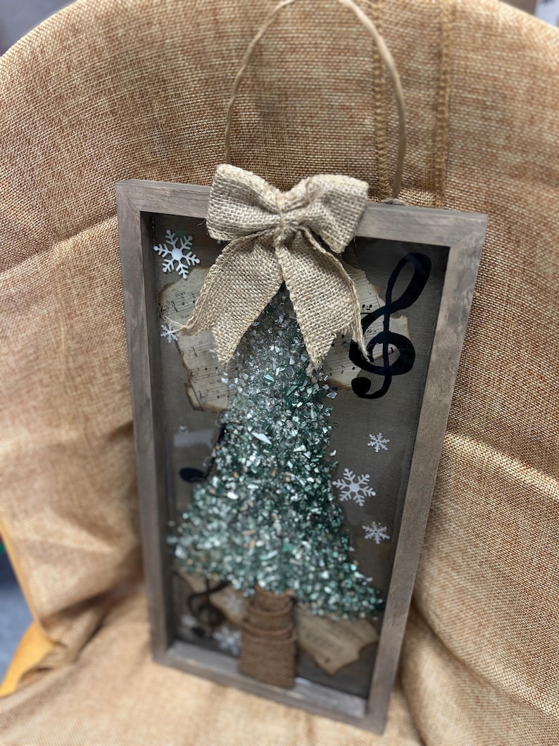 K&ouml;nnte beinhalten: Ein rustikaler Holzrahmen mit einer Schleife aus Sackleinen und einem gr&uuml;nen Glitzer-Weihnachtsbaum im Inneren. Der Rahmen hat eine Glasfront und ist mit Noten und Schneeflocken verziert.