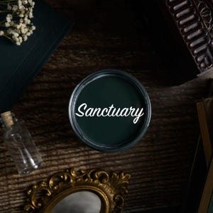May include: A circular container of dark green paint with the word Sanctuary written in white script on the lid, surrounded by a miniature Eiffel Tower, a small glass bottle with a cork stopper, a gold ornate frame, and several dark books.