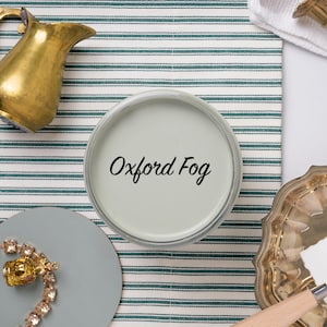 May include: A paint can labeled "Oxford Fog" sits on a striped fabric, surrounded by a gold pitcher, jewelry, and a palette knife. The color is a muted, light gray-green. The scene suggests interior design or home decor.