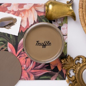 May include: A top-down shot features a can of "Truffle" paint, a color swatch, and a palette knife with paint. The background includes floral patterned paper, a brass pitcher, a woven basket, and a decorative gold frame.