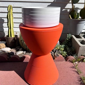 May include: A stack of white plastic plant saucers sits on top of a bright orange plastic stool. The stool has a wide, round top and a narrow, cone-shaped base.