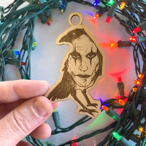 May include: Wooden ornament featuring a detailed laser-etched image of a character with dark eye makeup and a crow-like silhouette. The ornament has a hole for hanging and is surrounded by Christmas lights.